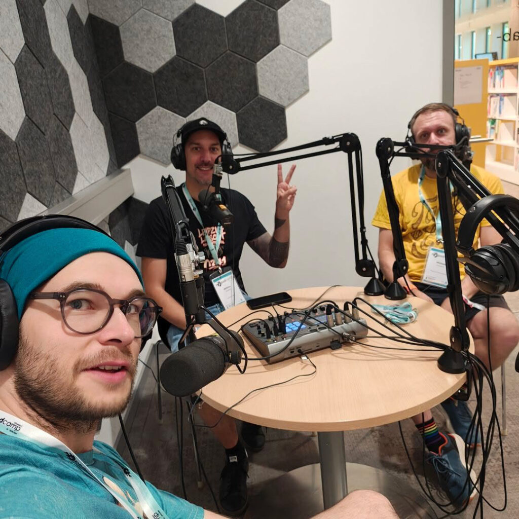 Drei Männer sitzen dicht beieinander in einem kleinen Podcast-Studio. Vor ihnen stehen mehrere Mikrofone an schwarzen Armen, dazwischen liegt ein kleines Mischpult auf einem runden Tisch, und ein Gewirr von Kabeln zieht sich über die Oberfläche. Links schaut ein Mann mit Brille und türkisfarbener Mütze direkt in die Kamera, in der Mitte lächelt einer und hält zwei Finger zum Peace-Zeichen hoch, rechts sitzt ein Mann im gelben T-Shirt ruhig neben seinem Mikrofon. Die Wände sind mit grauen, sechseckigen Akustikplatten verkleidet, was dem Raum eine sachliche, aber gemütliche Studioatmosphäre gibt.