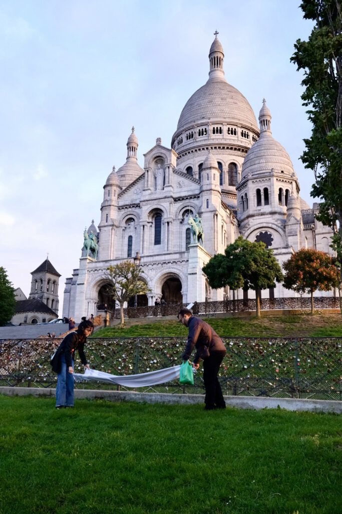ild im Hochformat

Die erhebt sich eindrucksvoll über dem Pariser Stadtteil Montmartre. 

Man blickt von einer grünen Wiese hinauf zu einer gewaltigen, hellen Kirche, die auf einem Hügel thront. Die weiße Basilika Sacré-Cœur Sie wirkt wie ein großes Schloss aus weißem Stein: In der Mitte steigt eine breite, runde Kuppel empor, darüber sitzt ein schmaler Turm mit einem Kreuz an der Spitze. Rechts und links stehen kleinere, ebenfalls rund geschwungene Türme. Die Fassade ist voller Bögen, Säulen, Fenster und steinerner Figuren – alles wirkt fest, kühl und sorgfältig gearbeitet.
Am Fuß der Kirche verläuft ein Zaun, dicht behängt mit vielen kleinen Liebesschlössern aus Metall. Davor, auf dem weichen Rasen, stehen zwei Menschen einander gegenüber. Sie halten gemeinsam ein langes, weißes Tuch zwischen sich, als wollten sie es ausbreiten oder zusammenlegen. Einer trägt dunkle Kleidung und beugt sich nach vorn, die andere Person steht links und zieht das Tuch zu sich.
Rechts rahmen hohe, dicht belaubte Bäume die Szene ein. Über allem liegt ein heller Abendhimmel. Das Bild verbindet etwas Großes und Feierliches – die mächtige Kirche – mit einem ganz einfachen, stillen Moment auf einer Wiese.