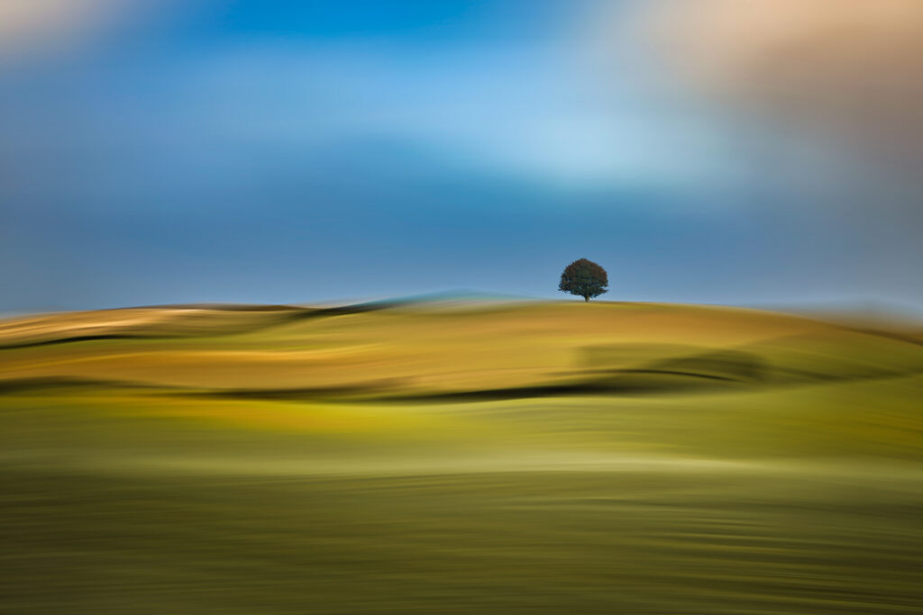 Bild im Querformat
Eine weite Landschaft in sanften Gelb- und Grüntönen zieht sich unter einem weichen, blauen Himmel dahin. Die Szene ist durch die Bewegung stark verwischt, sodass Hügel und Felder wie fließende Farbbänder wirken, fast wie ein gemaltes Bild. Der Himmel in der oberen Bildhälfte ist blau mit leichten weissen Bereichen.
Nur ein einzelner Baum am Horizont bleibt klar stehen und setzt einen ruhigen, kleinen Fixpunkt in diese sonst bewegte, fast träumerische Landschaft.