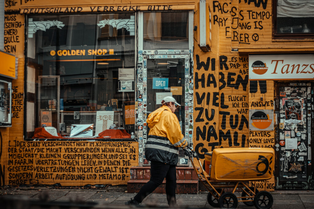 Bild im Querformat Hier sieht man die bunte Fassade der linken Buchhandlung Golden Shop in Bremen. Die Hauptfarbe des Bildes ist Gelb Die gelb-orangene Wand ist vollgeschrieben mit handgeschriebenen Sprüchen wie über dem Fenster“Deutschland verrecke Bitte’”, rechts daneben “Wir haben viel zu tun nämliches Geiles Life” und unter dem Fenster “Der Staat Verschwindet nicht im Kommunismus” und weitere 6 Zeilen Text. Rechts ist angeschnitten noch der Eingang einer Tanzschule erkennbar mit einer vol mit Aufkleber verklebten schwarzen Eingangstür Vor dem Laden schiebt ein Postbote in gelbem Regenmantel und Cap einen gelben Post-Bollerwagen. Auf dem Wagen ist vorne deutlich das Posthrin erkennbar. Der Postbote träge eine Türkise Bille. Die Fenster zeigen Bücherregale und eine beleuchtetes Schild mit dem Schriftzug “Golden Shop” – ein Bild voller Street-Art-Vibes.