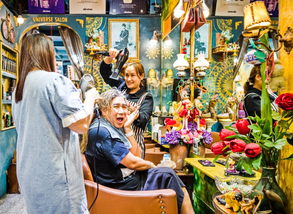 Bild im Querformat. Ein herrlich wuseliger, bis unter die Decke vollgestopfter Friseursalon, der aussieht wie eine bunte Schatzkammer. Die Wände sind tiefblau, verziert mit goldenen Mandalas, alten Gold gerahmten Schwarz-Weiß-Fotos und einem Meer aus Spiegeln, Lämpchen und Nippes – von Plastikrosen über Puppen bis hin zu goldenen Buddha-Figuren. 
Drei Personen sind zu sehen. Mittendrin sitzt ein Kunde mit nassen Haaren im Frisierstuhl, das Gesicht zu einer Grimasse der reinen Überraschung oder aus Spaß verzogen, während gleich zwei Frauen gleichzeitig mit Föhnen auf ihn zielen. Die Szene ist laut, bunt und wirkt wie ein wunderbar chaotischer, fast komödiantischer Schnappschuss aus dem prallen Leben.