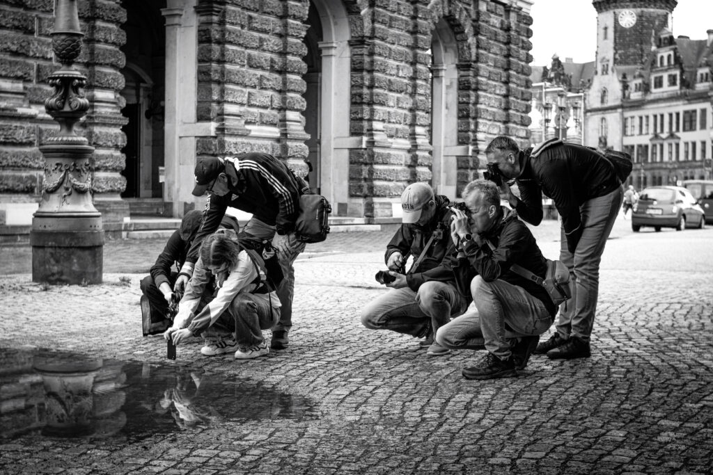 Schwarz-Weiss Bild im Querformat

Hier sieht man eine Gruppe von etwa sechs Fotografen vor einem historischen Gebäude.
Sie knien und stehen um eine große Pfütze auf dem Kopfsteinpflaster, halten konzentriert ihre Kamera und Handy hier rein. Jeder hat eine andere Position
Jeder hat Kamera und Stativ dabei, einige mit Rucksäcken und Caps.
Die Pfütze spiegelt das Gebäude mit Säulen, Statue und Turm perfekt wider.
In Schwarz-Weiß wirkt die Szene zeitlos und humorvoll.
Ein Moment der Leidenschaft, bei dem die Fotografen das perfekte Spiegelbild jagen.