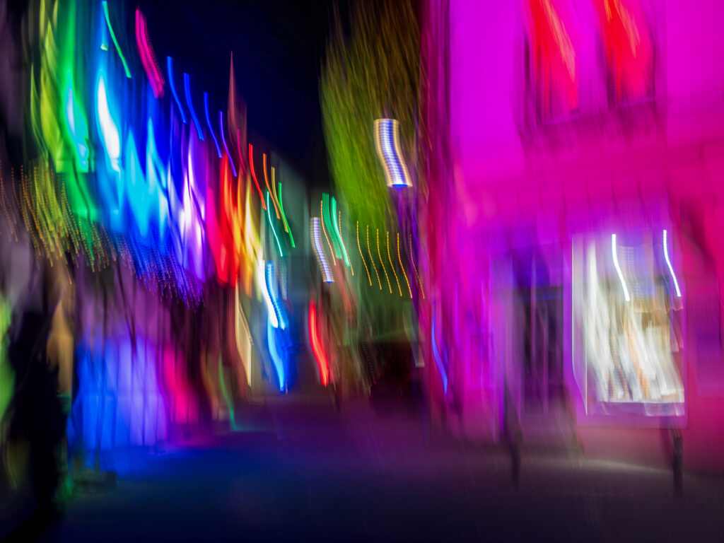Bild im Querformat
Hier sieht man eine nächtliche Straße mit Neonlichtern in voller Bewegung.
Bunte Lichtstreifen in Rot, Grün, Blau und Pink ziehen wie Regenbogenbahnen quer durchs Bild, als wären Autos mit langen Belichtungszeiten vorbeigefahren.
Die Lichter wirbeln und wellen sich, bilden einen psychedelischen Tunnel.
Gebäude und Fassaden verschwimmen zu pinken und lila Blöcken, mit scharfen Neonröhren als Ankerpunkten.
Die Szene pulsiert vor Energie, wie eine abstrakte Stadtparty in Farbe und Geschwindigkeit.