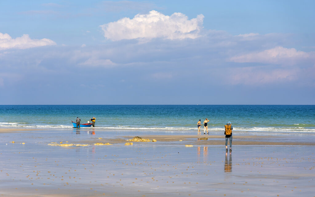 Bild im Querformat 

Ein weiter Strand in der Normandie liegt unter einem hohen, hellen Himmel mit großen, weichen Wolken. Das Meer schimmert in mehreren Blautönen, vom tiefen Dunkelblau am Horizont bis zum klaren Türkis näher am Ufer. Der nasse Sand spiegelt Himmel, Wasser und die kleinen Figuren am Strand wie ein glatter Spiegel. Vereinzelt stehen Menschen weit auseinander: einige am Ufer, andere mit Blick aufs Meer, und links zieht ein kleines Boot in der flachen Brandung die Aufmerksamkeit auf sich. Die Szene wirkt ruhig, offen und von dieser typischen nordfranzösischen Weite geprägt.